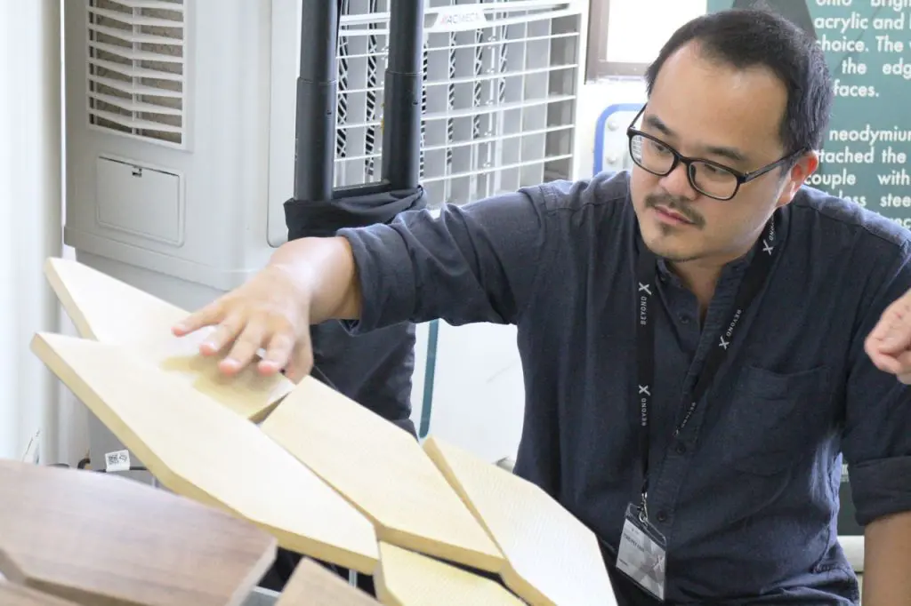A man wearing glasses and a dark shirt examines several light-colored, geometric wooden panels indoors near an air conditioner, possibly arranging components for a kinetic ceiling installation.
