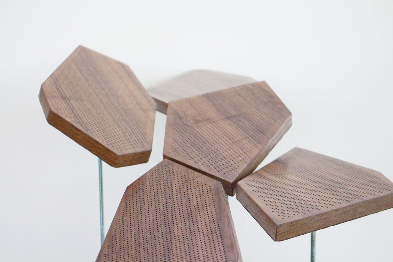 Five wooden, geometric-shaped panels with perforated surfaces are grouped together, supported by vertical metal rods against a plain white background, evoking the dynamic movement of a kinetic ceiling installation.