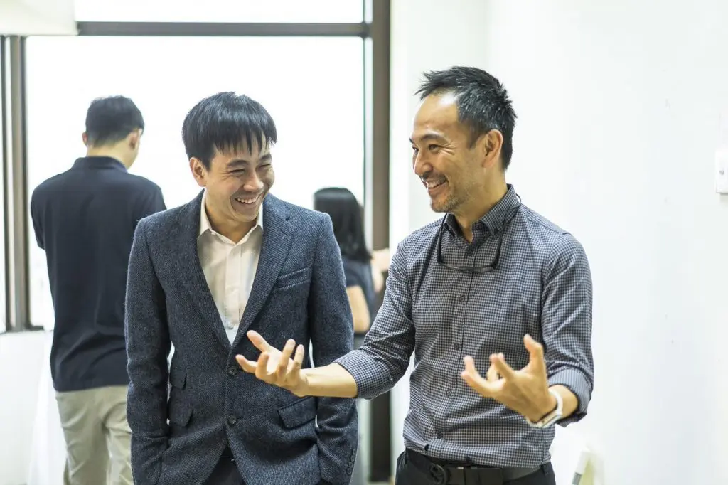 Two men stand indoors beneath a Kinetic Ceiling, smiling and conversing enthusiastically. One gestures animatedly with his hands, while the other listens, smiling. In the background, two more people face away near a large window.