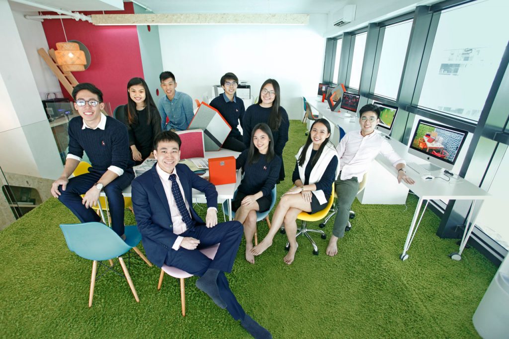 A group of nine young adults, dressed in business casual attire, pose and smile in a modern, brightly-lit office with green carpet, colorful chairs, and large windows. Some are seated while others stand behind them.