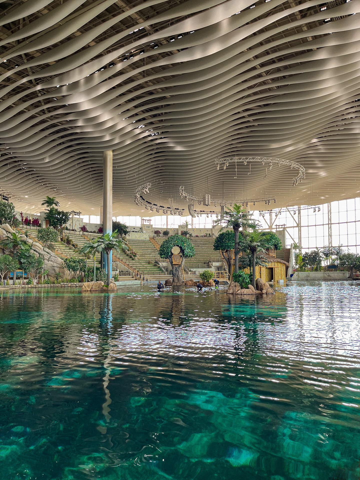 A large indoor water park with clear blue water, tropical plants, small huts, and modern wavy ceiling panels inspired by SeaWorld Abu Dhabi parametric facades. Sunlight streams through tall glass walls, creating a bright and inviting atmosphere.