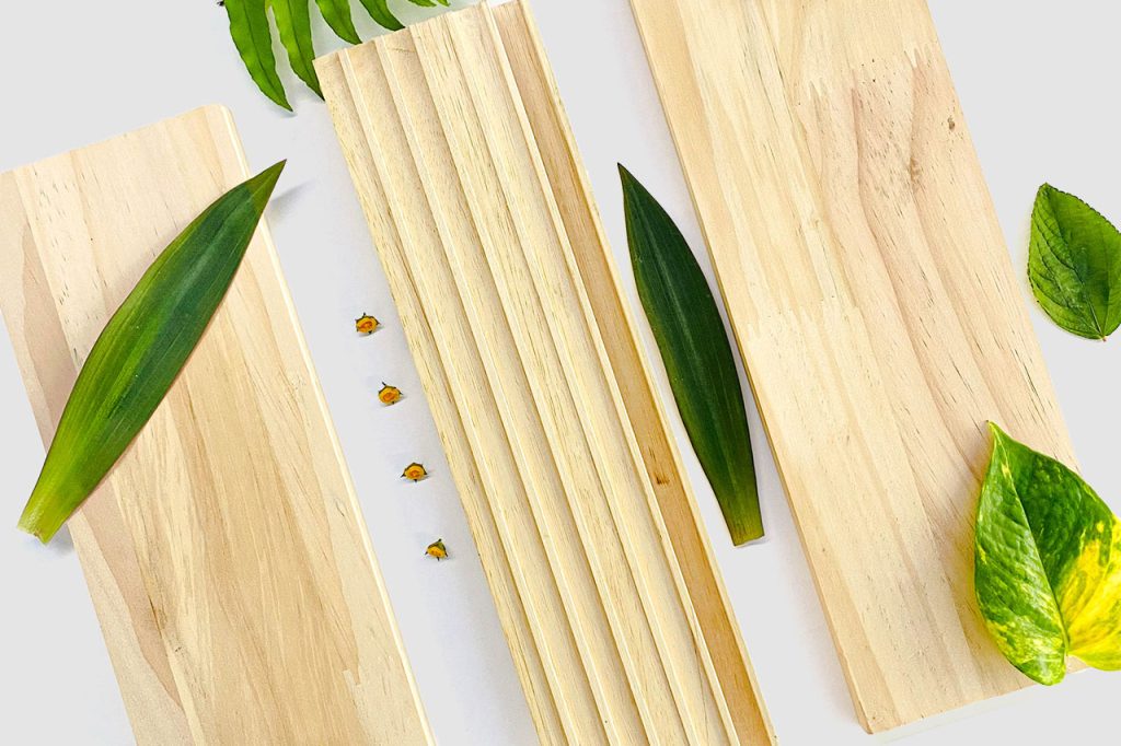 Three rectangular wooden boards arranged on a light surface, each decorated with different green leaves. Small yellow flower buds are placed between the boards, and additional green leaves are visible around the edges.